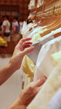 Woman hand touching and choosing light clothes hanging on wooden rack at fashion boutique store. Close up of female customer browsing light dresses and shirts, retail shopping consumer concept.