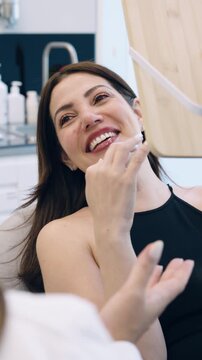 Satisfied female patient looking in the mirror, admiring her beautiful white teeth after a dental procedure at a modern aesthetics clinic. Vertical