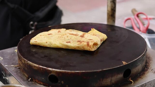 Vendor hand using T-shaped metal spreader to evenly distribute batter on hot circular griddle at street market in Nha Trang, Vietnam. Making thin crepe pancake, local fast food concept.