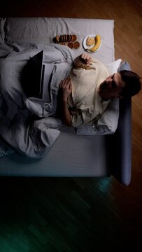 Man lying comfortably in bed during the night, consuming cookies and fruit while watching content on a laptop, depicting a scene of relaxation and unwinding at home