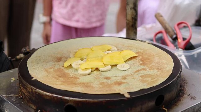 Vendor cooking sweet crepe with banana, mango, strawberry, condensed milk and chocolate sauce on hot griddle at street market in Nha Trang, Vietnam. Making fruit pancake, dessert concept.