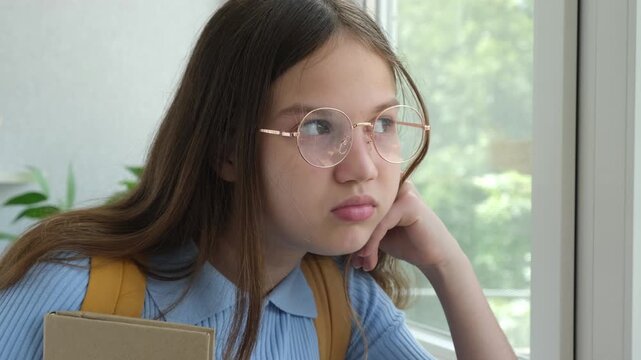 Thoughtful schoolgirl in glasses with backpack and book looking out of window. Unhappy teen girl alone