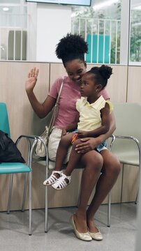 Young black woman sitting with her little girl on her lap in a hospital waiting area. Loving mother comforting her child before a visit. Vertical