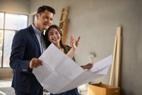 Happy couple talking while reading housing plans at construction site.