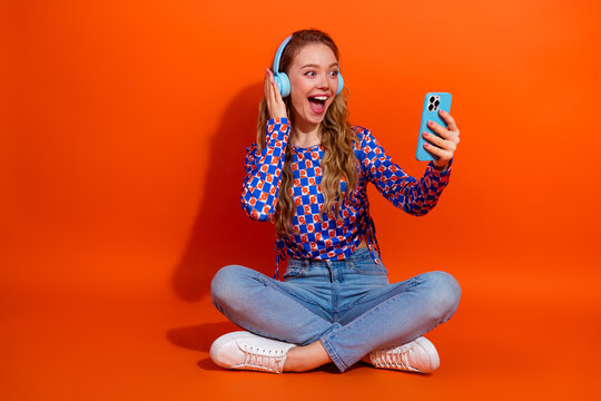 Cheerful young woman enjoying music with headphones while taking a selfie using smartphone against vibrant orange wall