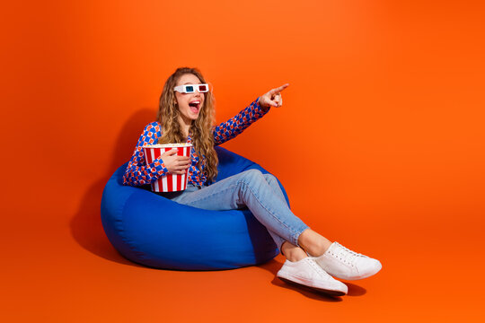 Young woman enjoying entertainment in 3D glasses while sitting on a beanbag chair with popcorn on an orange background.