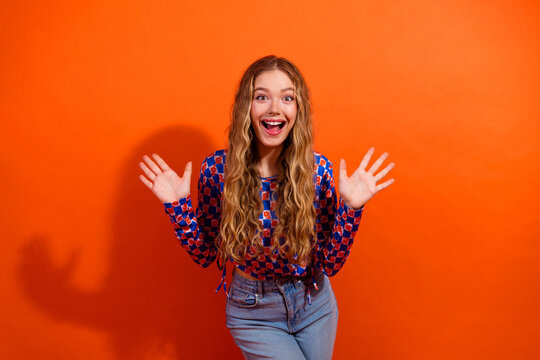 Cheerful young woman expressing excitement with hands raised in colorful casual outfit on vibrant orange background