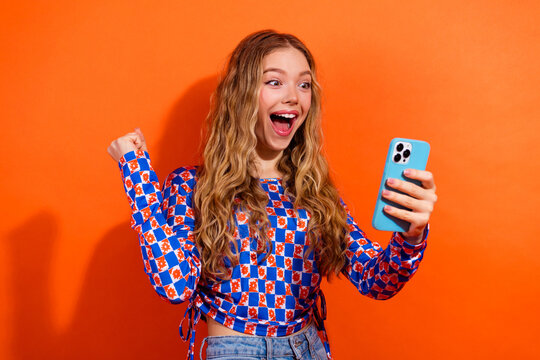 Excited woman in patterned top holding phone against vibrant orange background showcasing youthful joy and cheerful energy