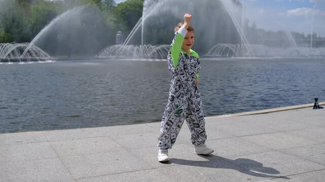 Energetic young boy in fashionable printed jumpsuit dancing dynamic modern street dance moves outdoors near river embankment with water fountains.