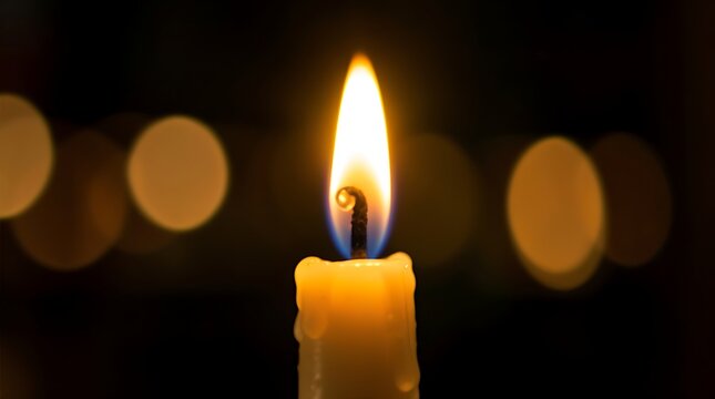 Lit candle wick with glowing orange flame against blurred warm bokeh lights in dark background closeup.