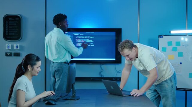 Young diverse IT team collaborating in a modern tech office. Software engineers coding on computers and discussing data on large screen late at night. Teamwork in software development and programming