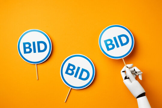 A white robotic hand holding a blue bidding paddle with "BID" signs among two others on a bright yellow background. Concept of artificial intelligence in auction and online bidding. Top view with flat