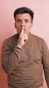 Serious middle aged man making a shushing gesture. He is putting his index finger to his lips, asking for silence against a pink background. Vertical