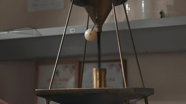 Gloved hand adjusts suspended off white test sphere beneath conical metal funnel in laboratory tripod setup with coiled wire support, precision fixture, warm diffuse lighting