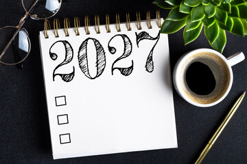 Naklejka premium 2027 New year resolutions on desk. 2027 goals list with notebook, coffee cup, plant on black table. Resolutions, plan, goals, action, checklist, idea concept. New Year 2027 resolutions. Copy space