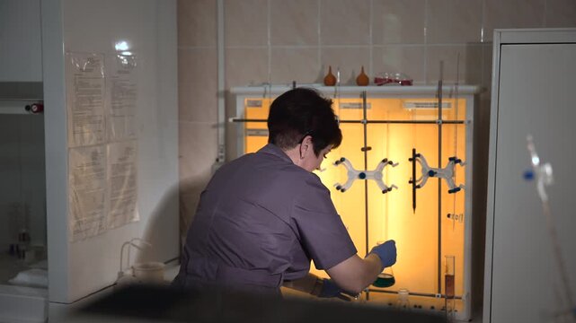 Laboratory technician handling glass flasks inside illuminated fume hood while comparing colored liquids in tiled chemistry workspace with protective gloves and dark uniform