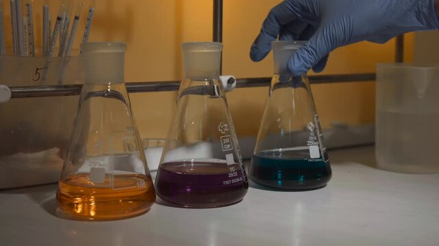 Gloved hand lifting blue green flask beside orange and purple flasks on laboratory bench under warm light, with sealed stoppers, clear glassware, and reflective liquid surfaces