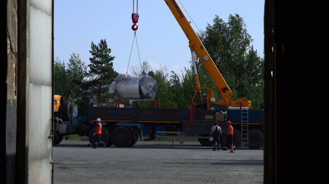 Mobile crane lowering wrapped cylindrical metal tank onto flatbed truck while ground crew guides careful placement through doorway framed industrial site view, with crane, lowering, tank, truck detail
