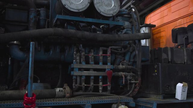 Industrial machinery compartment packed with hoses, pipes, valves, fittings, control boxes, worn metal surfaces, grime, and bright orange panel on heavy duty equipment
