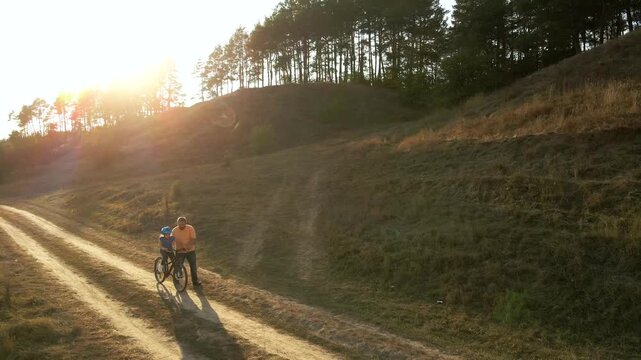 Wide angle view of a father helping his young daughter learn to ride a bicycle along a rural dirt road surrounded by hills and pine trees during warm golden hour sunset.