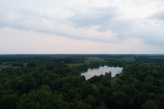 Aerial drone view over lush green trees and a calm lake under a soft twilight sky with clouds