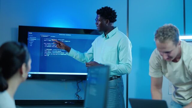 Diverse software development team brainstorming code in a modern dark office. African American tech lead presenting programming strategy on screen to colleagues during a late night startup meeting.