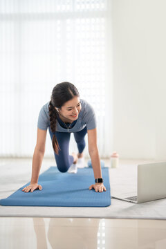 Young woman do excercise on a yoga mat at home, following a online workout tutorial on laptop.