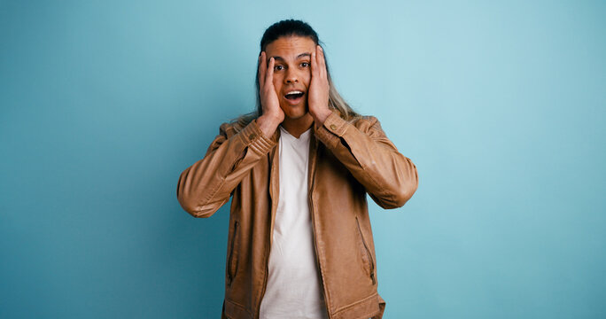 Young Latino man reacting with amazed, shocked expression, hands on face, looking at camera on blue studio background
