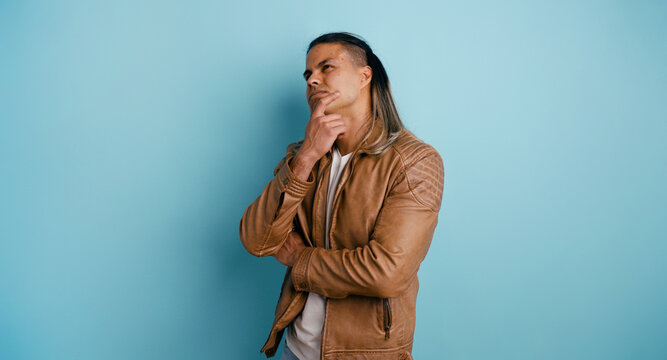Man in leather jacket thinking, looking up with hand on chin, brainstorming ideas, contemplating options on a blue background
