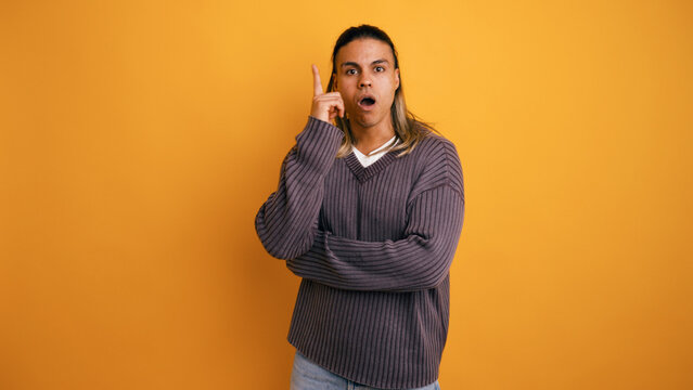 Young man with long hair pointing finger up, having a sudden realization or inspiration. Posing on yellow studio background