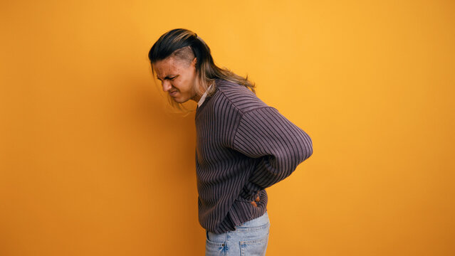 Young man experiencing intense back pain, holding his aching lumbar region with a grimacing expression on a yellow background