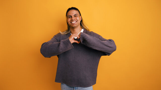Young latino man enjoying and expressing love, making a heart symbol with his hands standing in a studio with a yellow background
