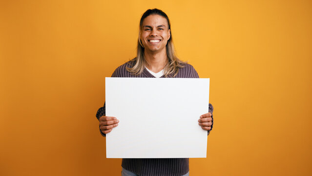 Smiling young Latino man presenting a blank white sign, offering a space for custom messages or promotions against a yellow background