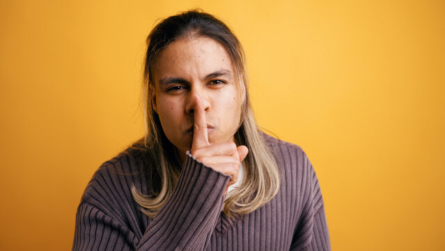 Young man standing in a studio, making a shushing gesture with his finger on his lips, emphasizing quiet and secrecy