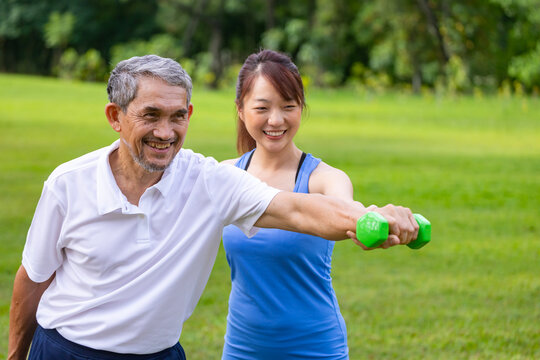 Senior asian man is using dumbbell for weight training strength while his daughter is supporting in the public park for elder longevity exercise and outdoor workout