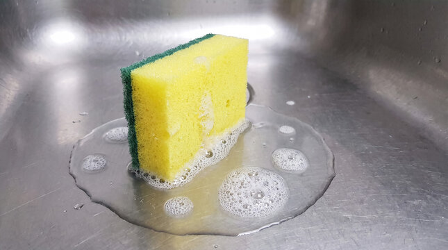 Yellow sponge scrubs dishes in a stainless steel sink, producing abundant soap suds and foam