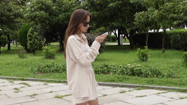 young woman checks a smartphone while walking through a green city park, representing outdoor navigation, travel planning and a connected summer lifestyle during independent travel in the city