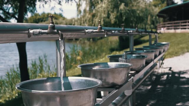 A row of stainless steel wash basins and brass faucets stands outdoors by a lake while water flows from one tap. Concept of outdoor hygiene, camping amenities, and summer recreation.