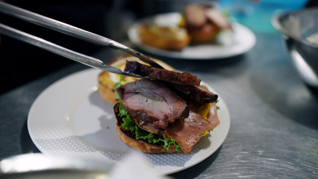 Chef using tongs to place slices of pork meat on a fresh sandwich bun with lettuce. Focusing on preparing delicious street food
