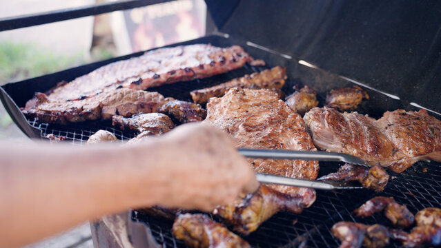 Man using tongs to turn various meats including ribs and chicken on an outdoor barbecue grill during a local gathering