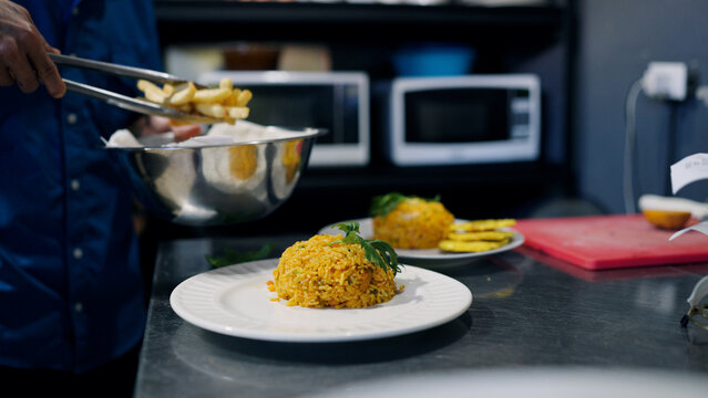 Chef preparing a meal in a professional kitchen, adding french fries to plates with yellow rice and plantains