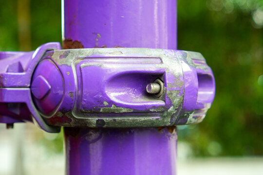 Close-up of a weathered purple industrial pipe clamp with visible rust and peeling paint, secured by a bolt against a blurred green background, showcasing mechanical wear and outdoor durability.