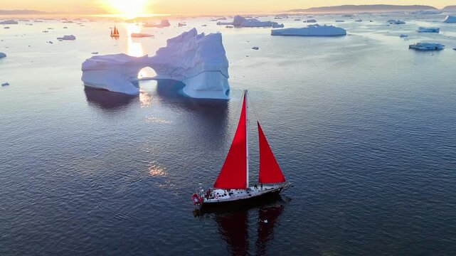 Red sailboats glide past a drifting Arctic ice floe with a natural arch, illuminated by golden sunset light. A stunning contrast highlighting the beauty and fragility of a changing polar world.
