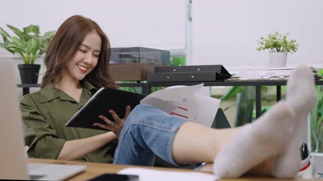 Happy beautiful Asian woman real estate business owner sitting smiling comfortable at her private desk while checking and analyzing histogram data from paperwork files at the workplace in the office. 