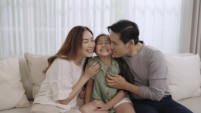 Happy Asian family, father, mother, and a child girl sitting on the sofa in the living room at home and kissing their daughter's cheek together with love and looking at the camera, Family Day Concept.