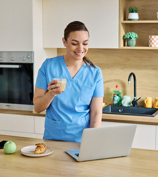 Portrait of a young nurse getting ready for work and using laptop online meeting standing in kitchen home. Overworked, busy, stress, urgency, multitasking concepts