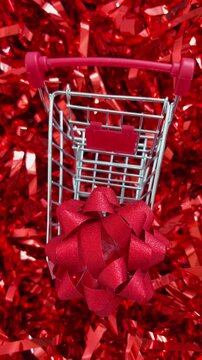 Miniature shopping cart with a red gift bow slowly moving over a shiny red tinsel background, symbolizing christmas holiday shopping, black friday sales, and consumerism