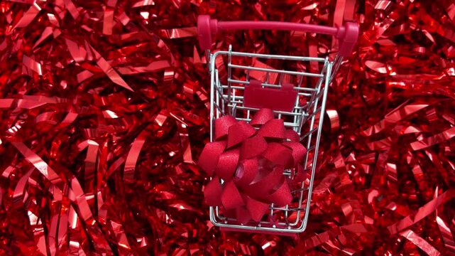 Shopping cart with red gift bow slowly moving on a shiny tinsel background representing holiday shopping, christmas sales, consumerism, and valentine's day presents