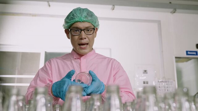 Professional Asian man inspector engineer in uniform standing holding an empty glass beverage bottle on a conveyor belt with explaining how to inspection by video call in the factory production line. 