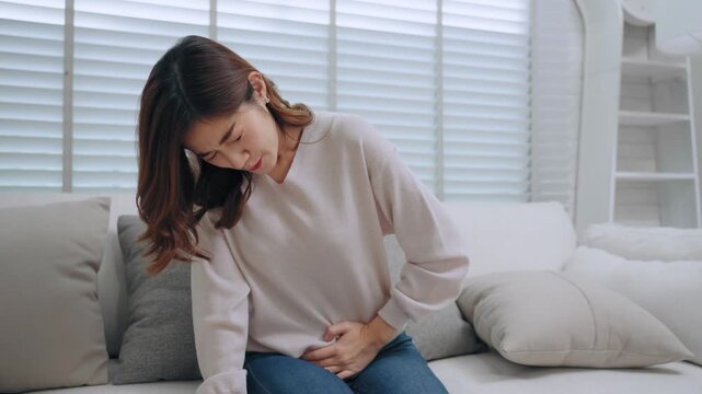 An Asian young woman sitting on sofa and holding her painful stomach, suffering from severe abdominal pain, menstrual cramps, food poisoning, indigestion, or gastritis symptom at home living room
